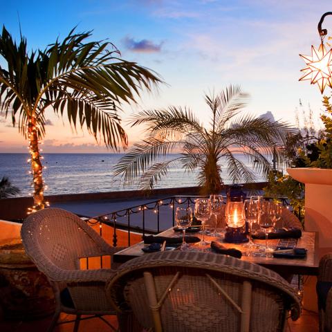 The image shows a picturesque outdoor dining setup on a balcony overlooking the sea at sunset