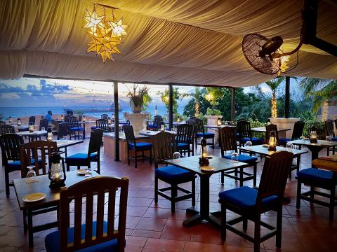 This image shows an outdoor dining area of a restaurant at sunset with a view of the ocean