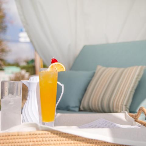 The image features a refreshing scene at an outdoor setting, where a glass of orange cocktail garnished with a slice of orange and a red cherry is placed on a wicker table