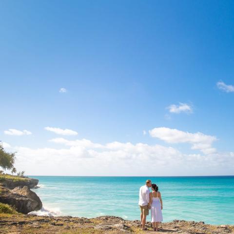 This image depicts a couple standing on a rocky coastline, sharing a kiss by a stunning turquoise ocean under a clear blue sky with a few clouds