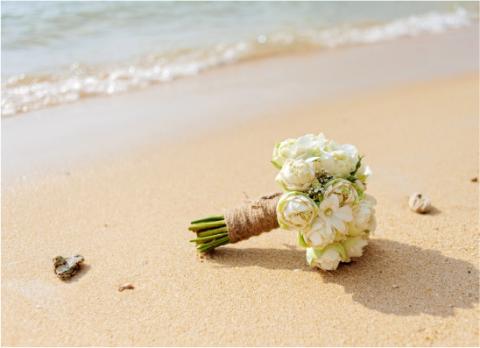 This image shows a bridal bouquet of white roses with some greenery and small flowers, tied with a twine-wrapped handle, lying on a sandy beach