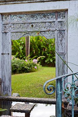 The image shows a beautifully designed garden scenery featuring an antique-looking gate with intricate metal designs and weathered wooden posts