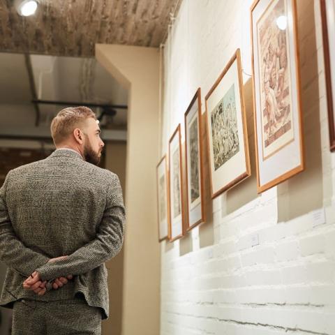 The image depicts a man viewing framed artworks hung on a white brick wall in an indoor gallery setting