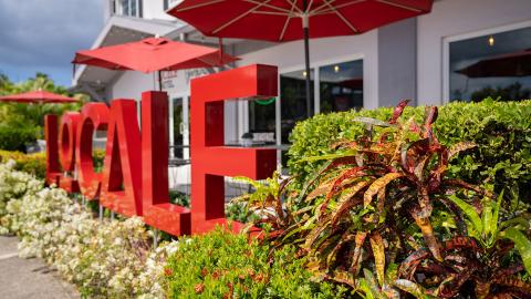 The image shows an outdoor setting with a large, red sculpture or installation spelling out the word "LOCAL"