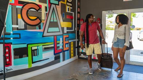 This image features a man and a woman entering a brightly lit room, possibly a gallery or a modern space, characterized by a colorful and abstract mural on the wall