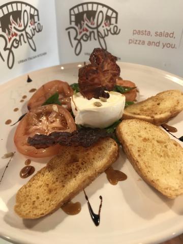 The image features a dish served on a white plate, which consists of toast slices, a portion of cheese (possibly mozzarella), slices of tomato, and green leafy vegetables possibly basil or spinach, topped with a piece of crispy bacon