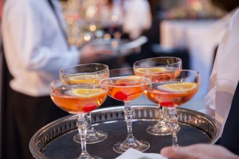 The image features a close-up view of a tray held by a server, containing several glasses filled with a vibrant orange-colored cocktail