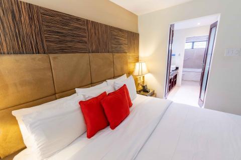 The image shows a well-lit modern bedroom, featuring a large bed with a plush upholstered headboard set against a decorative wood panel wall