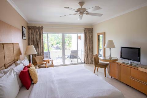 The image shows a well-appointed hotel room that features a large double bed with beige headboards and white bedding, complemented by contrasting colored pillows