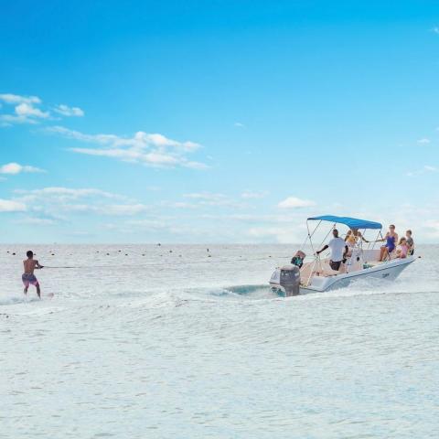 The image shows a scenic beach setting with a leisure boat moving towards the right, carrying several passengers, and a person water skiing to the left of the boat