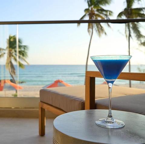 This image features a refreshing blue cocktail in a martini glass, placed on a round table at an outdoor beachside setting