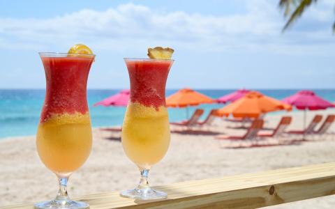 This image shows a tropical beach setting with two colorful cocktails placed on a wooden bar
