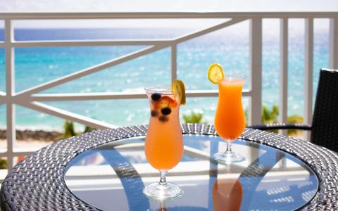 This image features a scenic view of a beach from a balcony, with two cocktails placed on a round glass table