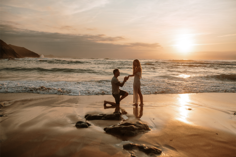 This image captures a romantic scene on a beach at sunset where a man is kneeling and holding a woman