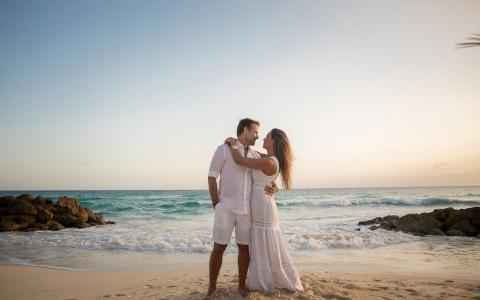 This image features a couple standing on a sandy beach at sunset, embracing and looking at each other affectionately