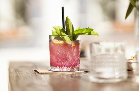 The image features a delicious-looking cocktail served in a textured glass, with a straw and garnished with a sprig of fresh basil and a slice of lemon