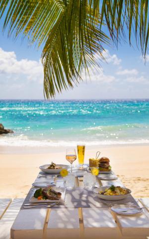 This image features a beautifully set dining table on a sandy beach, under the shade of palm leaves, facing a clear blue sea