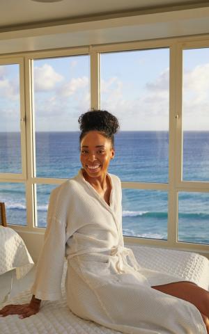 The image shows a woman sitting on a bed near a window, smiling at the camera