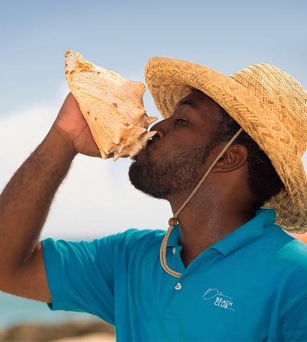 This image showcases a man wearing a straw hat, drinking or pretending to drink from a large conch shell