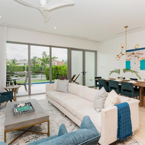 This image shows a modern, well-lit living room that seamlessly transitions into a dining area, all overlooking an outdoor patio with a pool