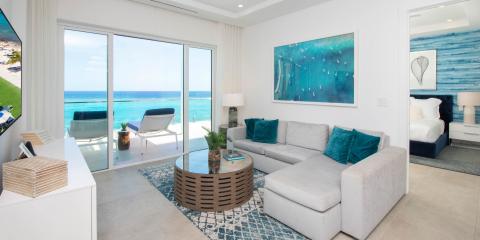 The image displays a well-furnished, modern beachfront apartment with a spacious living area that opens directly onto a sandy beach and a clear blue sea