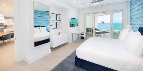 The image shows a modern beachfront bedroom that opens directly onto a balcony with a view of the sea
