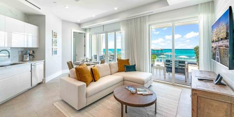 This image features a modern, coastal living room with large sliding glass doors that open to a terrace overlooking a breathtaking beach and turquoise ocean