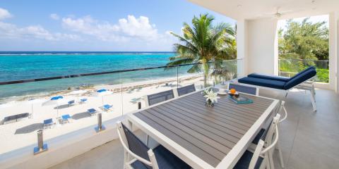 This image features a spacious, beachfront balcony with a stunning view of a turquoise ocean and a sandy beach lined with sun loungers and umbrellas