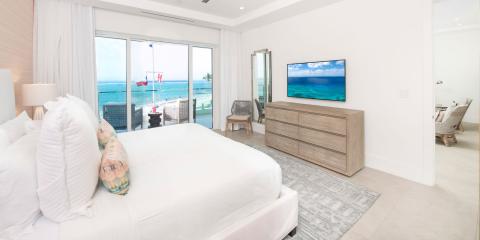 This image shows a bright and modern beachfront bedroom, with large sliding glass doors that open to a balcony overlooking a beautiful sandy beach and ocean