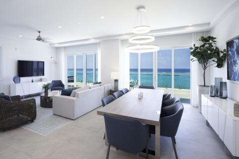 The image depicts a modern, bright beachfront living space with a clear view of the ocean through large glass doors