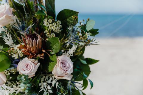 This image features a beautiful floral arrangement with a variety of flowers and foliage, set against a serene beachfront backdrop