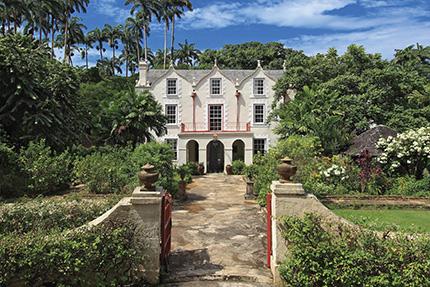 This image shows a grand, colonial-style mansion surrounded by lush greenery