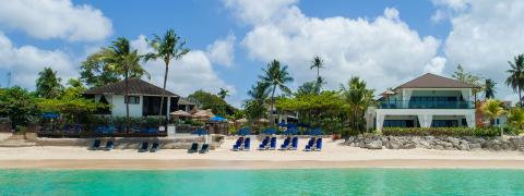 The image displays a serene beachfront view featuring a clean sandy beach, clear turquoise water, and a row of elegant vacation homes or resorts