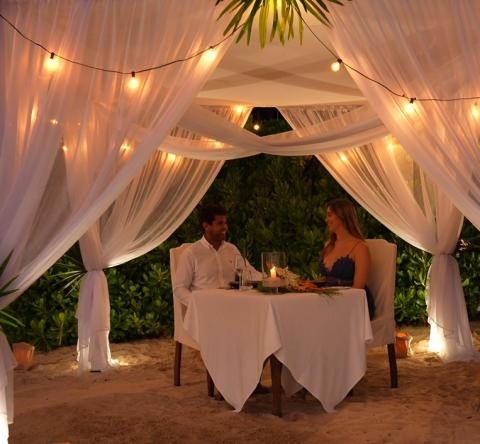 The image features a romantic dinner setting on a sandy area, likely a beach, under a canopy adorned with white drapes and string lights