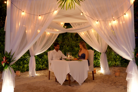 This image features a romantic beach dining setup with a couple sitting at a table under a beautifully decorated canopy with sheer white drapes and warm string lights