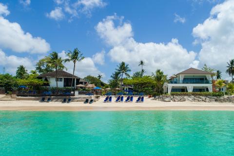 This image shows a tranquil beachfront scene with clear turquoise waters in the foreground and a sandy beach adorned with neatly arranged blue lounge chairs