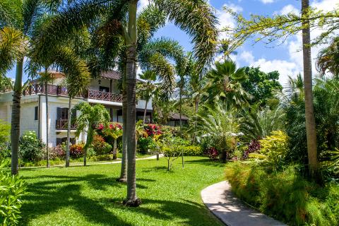 This image features a lush tropical garden surrounding a multi-story building with stone and wooden architecture