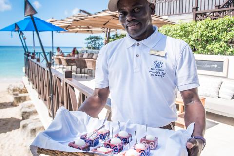 This image shows a man holding a basket filled with rolled towels, each decorated with a flower, likely serving them to guests at a beachside establishment