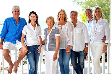 The image features a group of seven adults—four women and three men—posing together on what appears to be a beach house balcony