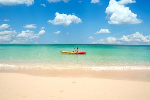This image captures a serene beach scene with a person kayaking on crystal-clear, shallow waters near the shore