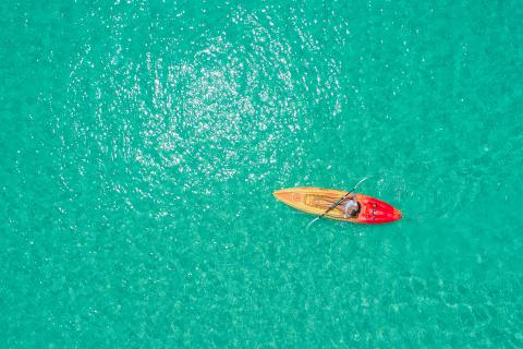 The image depicts an aerial view of a person in a red kayak paddling on a shimmering turquoise sea