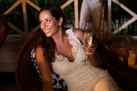 This image shows a woman sitting in a wicker chair on a balcony or patio, dressed in an elegant, lace-detailed dress, possibly a wedding gown