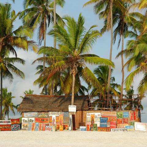 This image features a tropical beach setting with a small, rustic building adorned with colorful paintings and a thatched roof