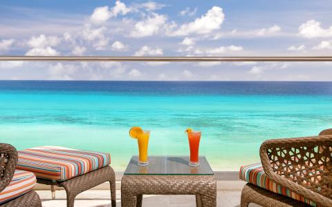 This image shows a beautiful beachfront setting with two colorful cocktails placed on a glass tabletop