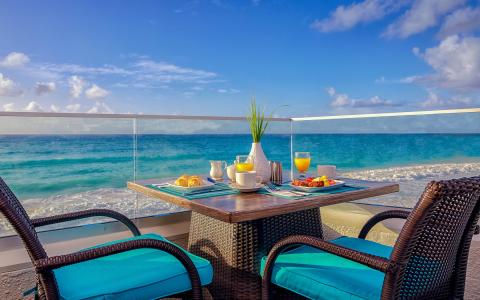 The image shows a beautifully set breakfast table on a balcony overlooking a beach