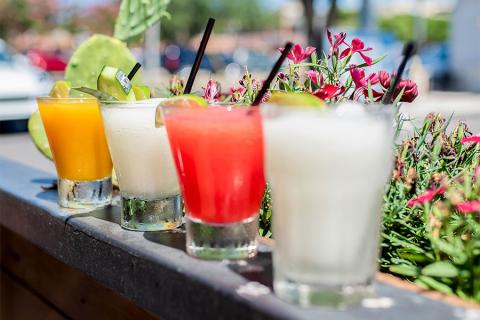 This image features a selection of colorful beverages arranged on a wooden ledge, possibly at an outdoor bar or restaurant setting