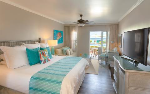 This image shows a well-decorated hotel room that features a large bed with white and turquoise linens and several pillows of varying sizes and colors