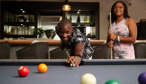 This image shows two people playing pool in an indoor setting that resembles a bar or lounge area