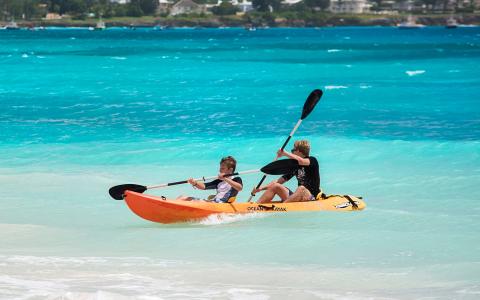 This image shows two people kayaking in a bright orange tandem kayak on clear turquoise ocean waters
