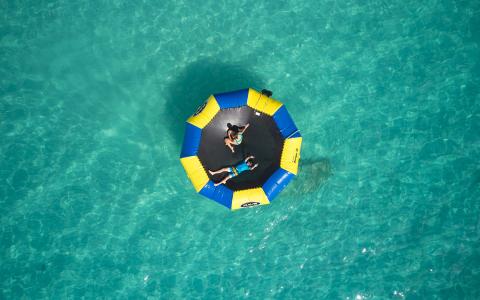 The image features two people lounging on a large, circular floating raft in a clear turquoise body of water